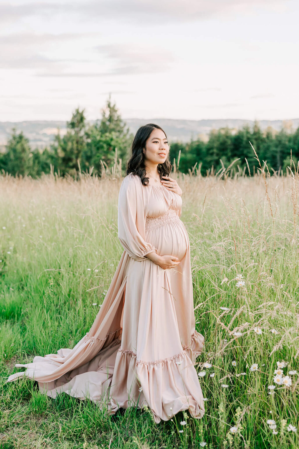 Portrait of a woman in a green field with white flowers. She is wearing a cream rose gold dress and hand a hand on her belly.