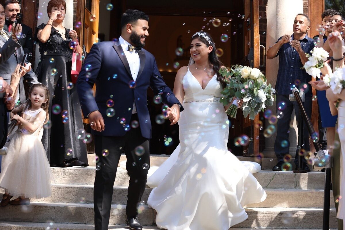 Bride and groom walk through bubbles outside church with guests cheering