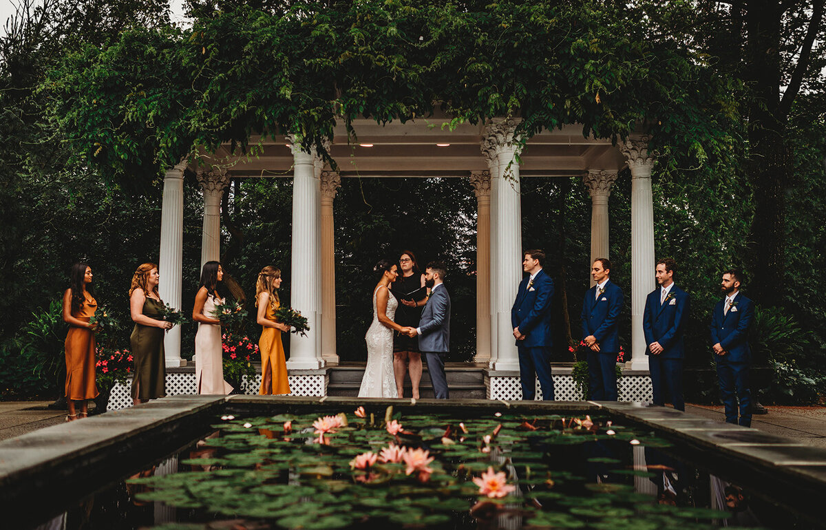 Outdoor courtyard wedding captured by Marilyn wedding photographers with bride and groom holding hands at the end of a reflecting pond and a pergola made of stone is behind them photographed by Maryland wedding photographer