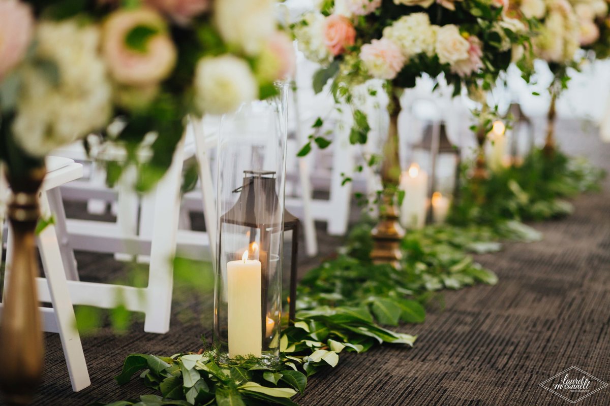 wedding aisle lined with greenery garlands, lanterns, and large flower arrangements on gold stands