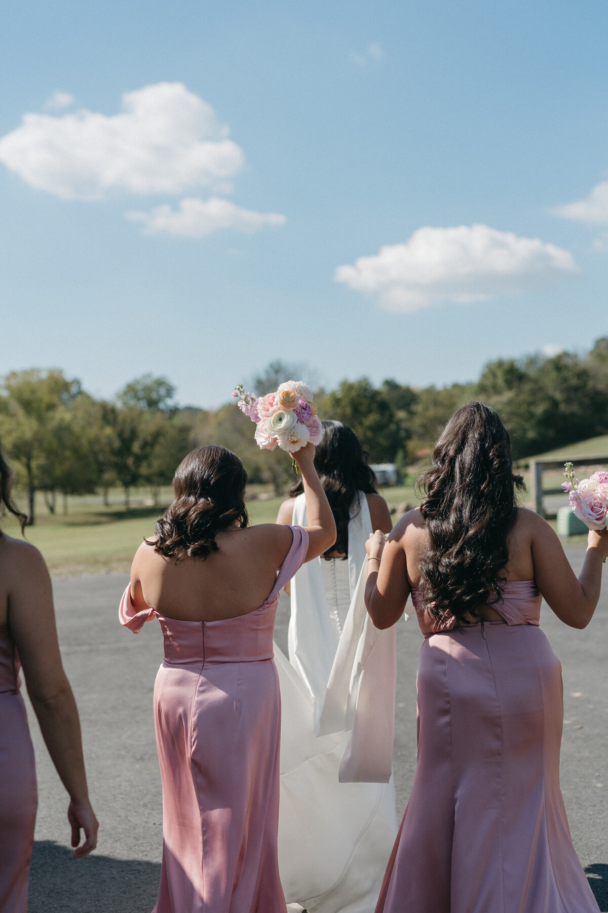 Bridesmaids in blush satin dresses carrying pastel bouquets of roses, dahlias, and ranunculus for a cohesive, romantic floral look as they walk toward an outdoor ceremony.
