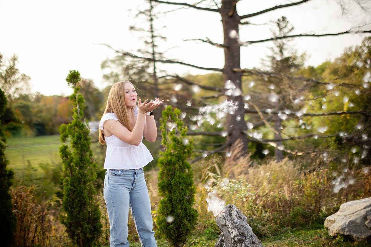 A teenage girl in a white top and jeans blowing dandelion seeds in a field, surrounded by trees and late afternoon sunlight, creating a magical and carefree scene in Metro Detroit. and Farmington Hills