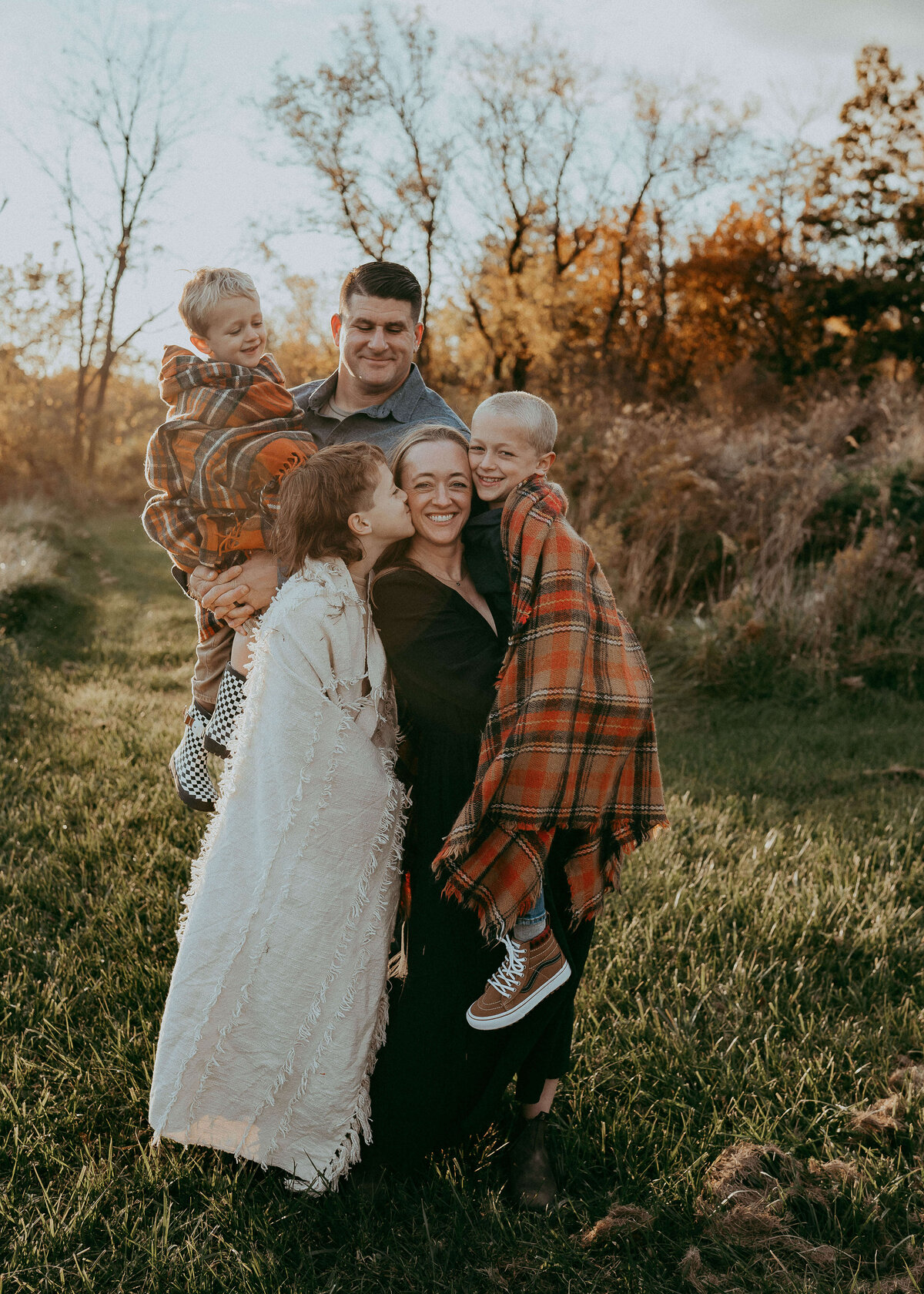 Parents cuddle with their three boys during a family photo session that takes place in fall so everyone is wrapped in blankets to stay cozy while the gorgeous sunset beautifully paints halos around their bodies.