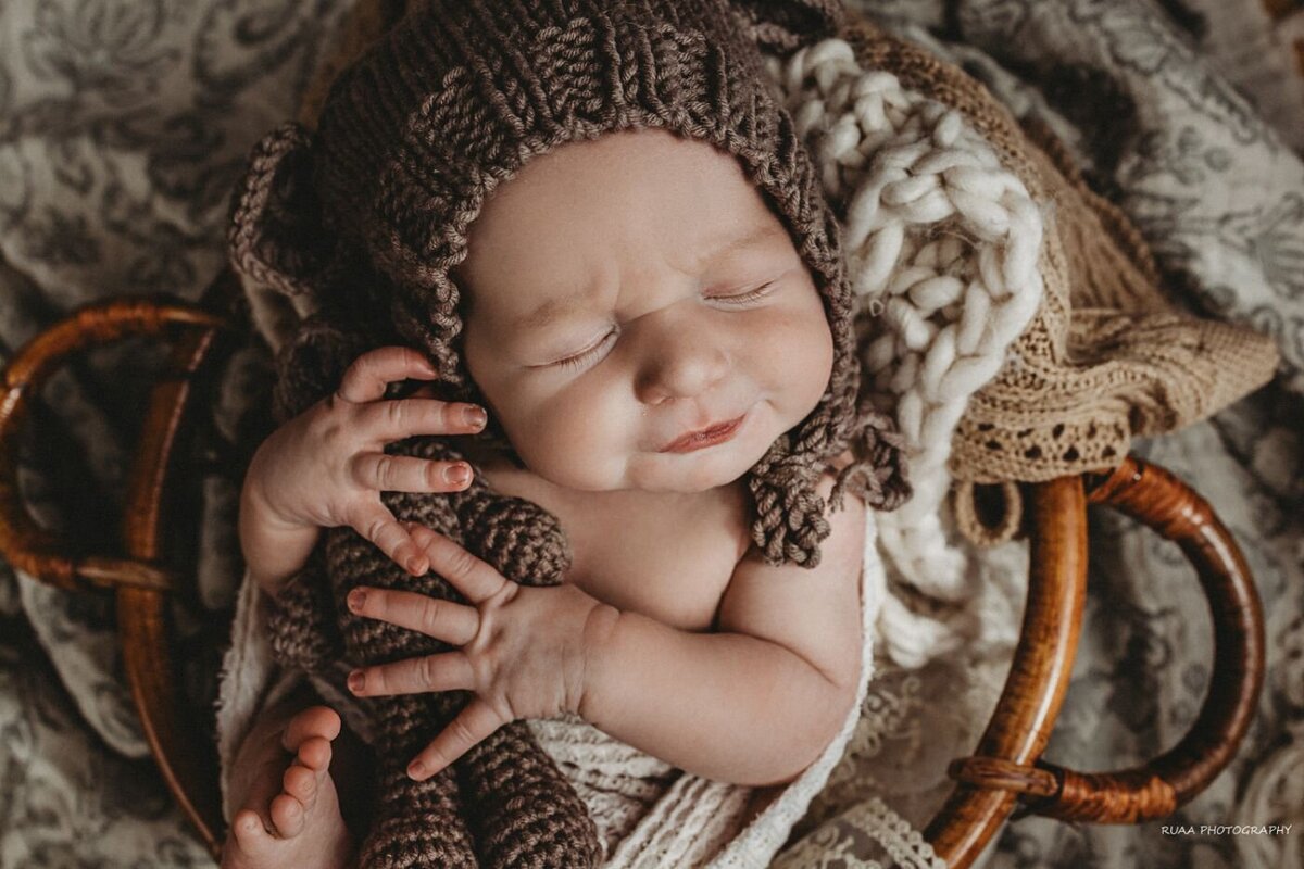 Serene close-up of a newborn baby in a deep sleep, wearing a charming brown textured bonnet and cuddling a tiny knitted bear while lying in a cozy basket