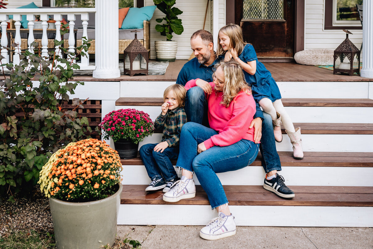 The family sits together on the front steps of their home. The mother, father, and children are all smiling and relaxed, surrounded by vibrant potted flowers.