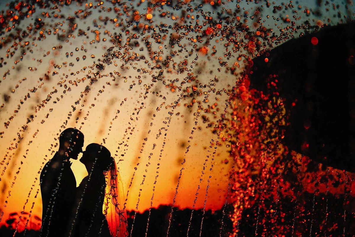Silhouette of a couple kissing behind a spray of water droplets with a vibrant orange sunset in the background