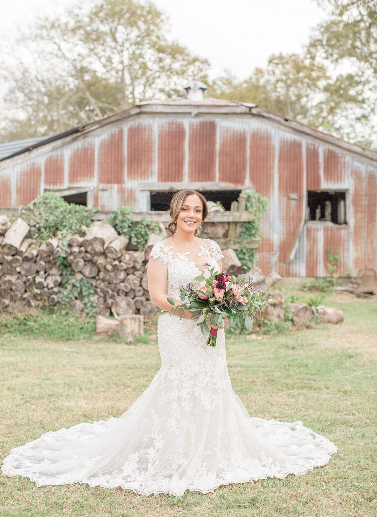 The bride stands in front of a rustic building holding her bouquet and smiling for the camera