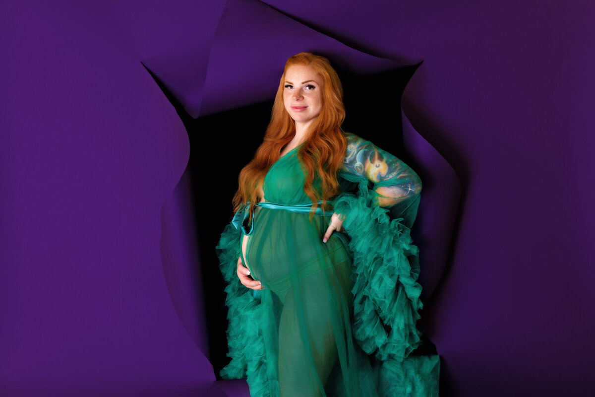 Maternity Photographer- a pregnant woman holds her belly in front of a purple backdrop