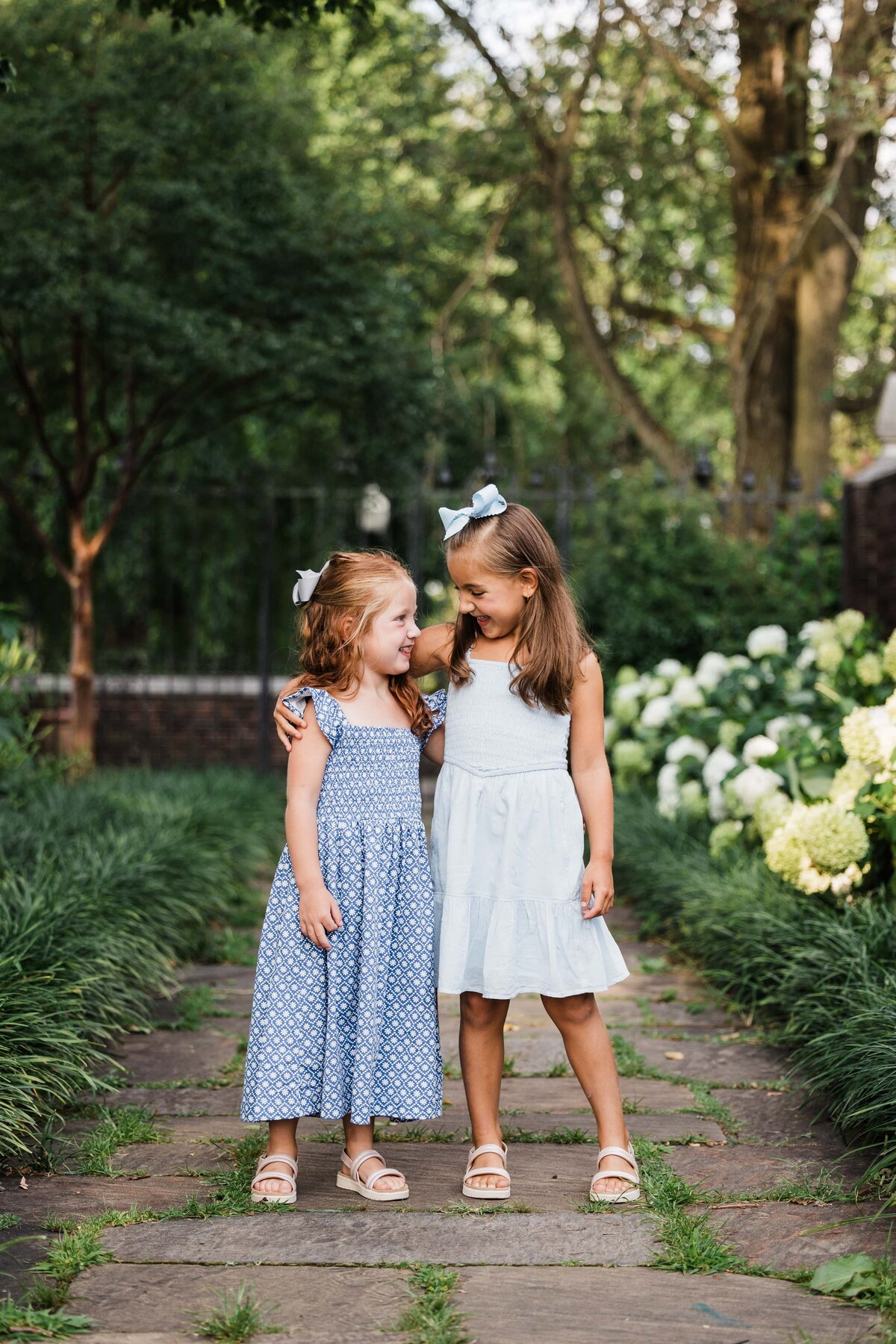 Two young girls smiling at each other on a garden path, captured beautifully by a family photographer in Pittsburgh.
