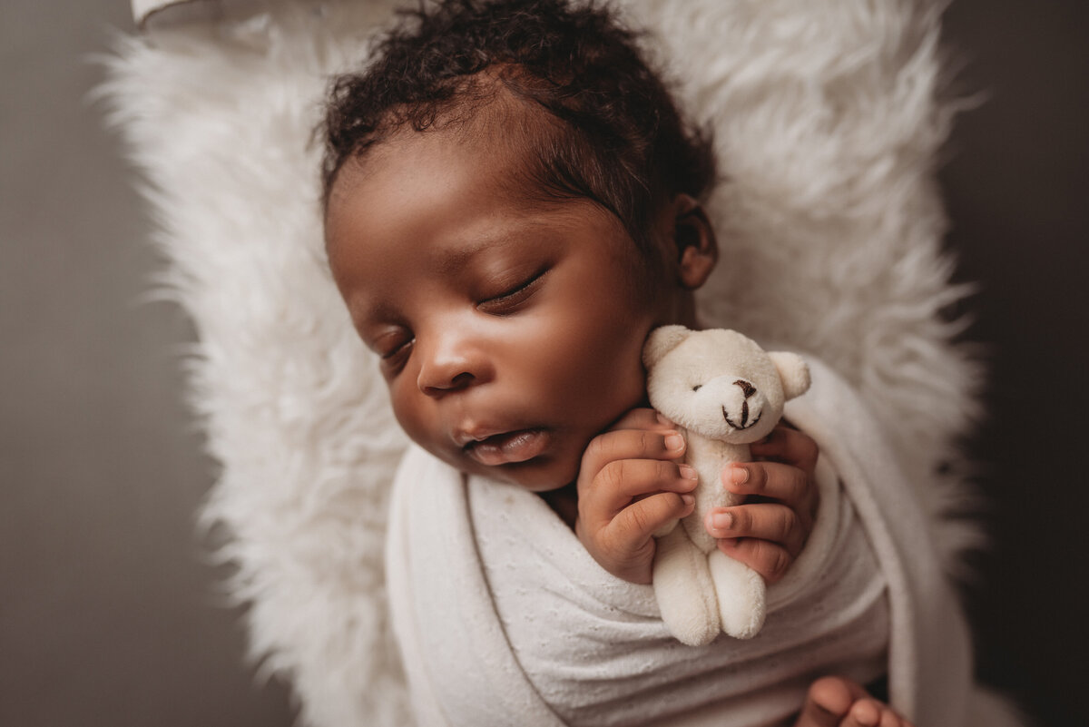 sleeping newborn boy with black curly head of hair wrapped in white swaddle laying on white fur blanket holding mini teddy bear