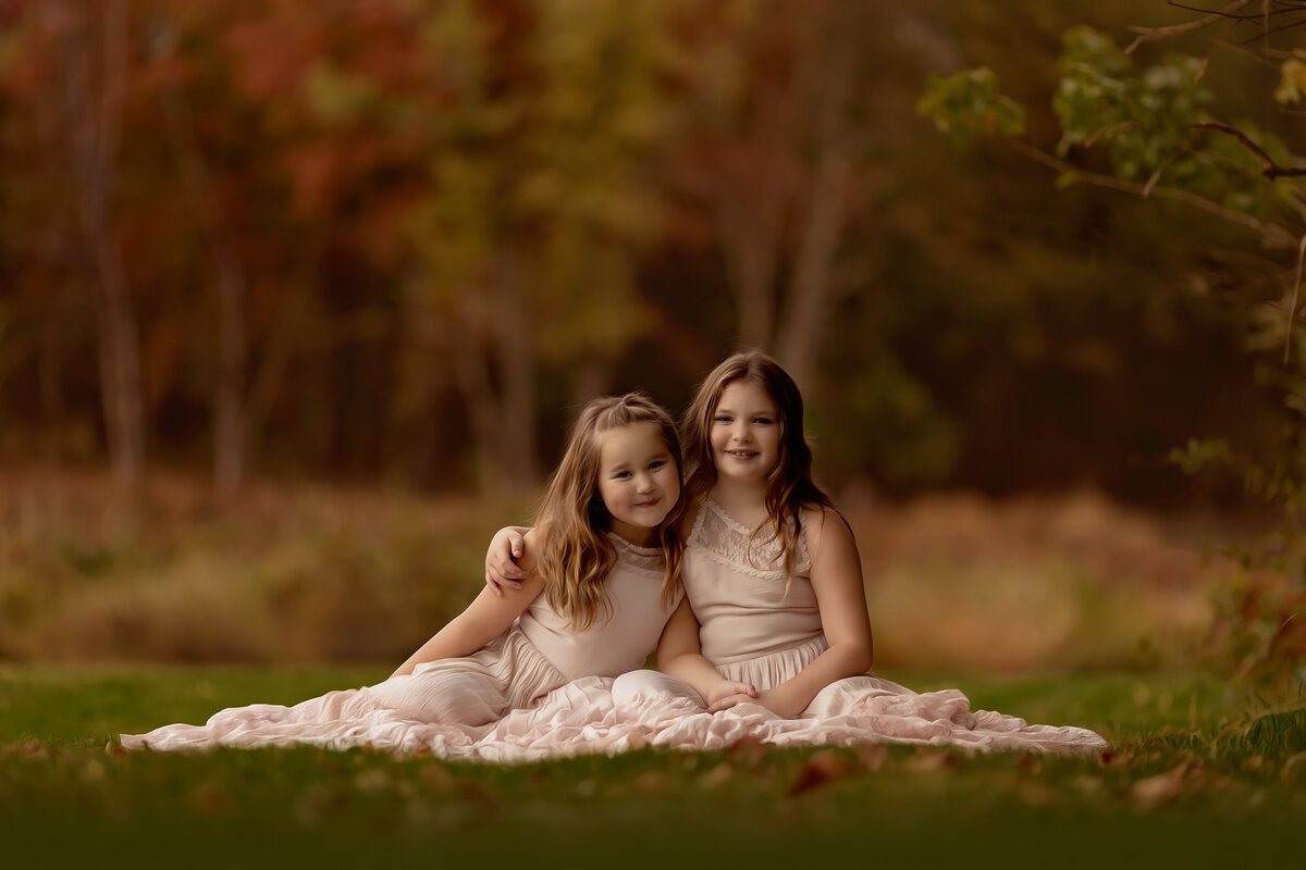 Two young sisters wearing identical dresses cuddle on a picnic blanket in a grassy lawn at sunset