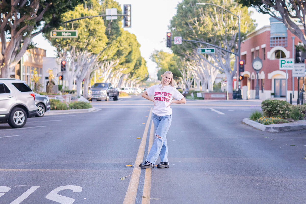 A person stands confidently in the middle of a sunny street, wearing a white shirt with red lettering and ripped jeans. Trees line the road, and buildings and cars are visible in the background.