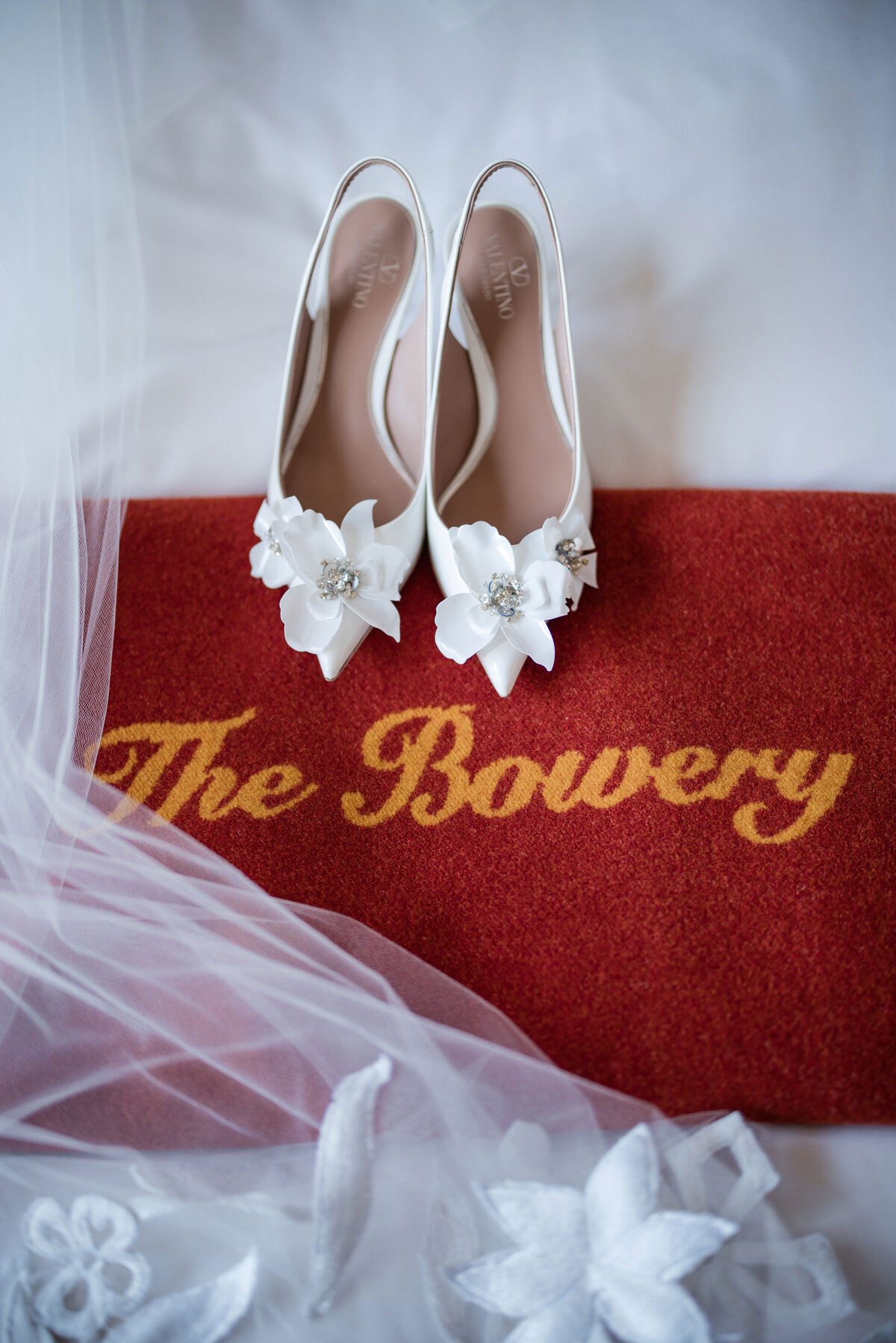 White floral shoes rest on a red mat with "The Bowery" written on it; sheer fabric is draped around the scene. This photo was taken by a luxury NYC wedding photographer specializing in intimate ceremonies
