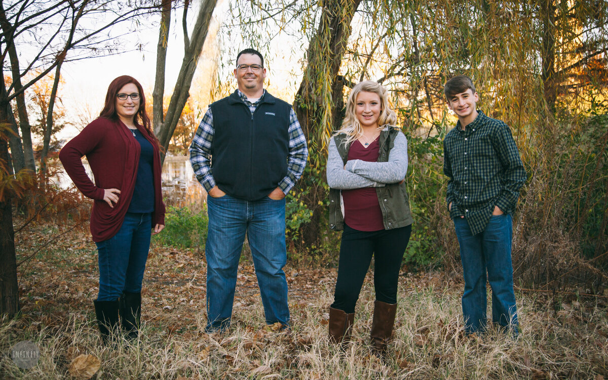 A happy family of four poses together in a scenic outdoor setting surrounded by autumn foliage. Dressed in coordinated fall outfits with warm tones, they stand in a natural wooded area with golden leaves and soft lighting. The parents and two teenagers exude warmth and connection, making this the perfect inspiration for fall family photography sessions.