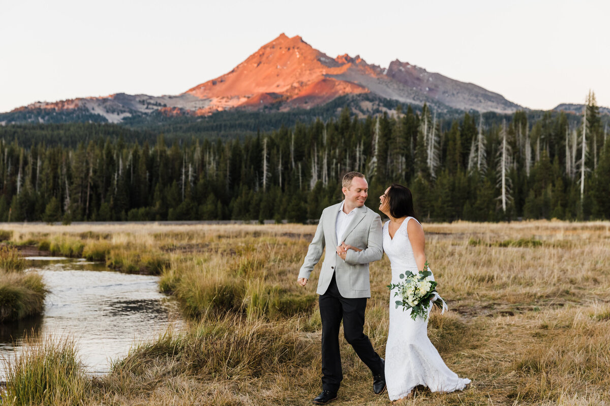 A bride and groom walk along a creek as the alpenglo lights up Broken Top Mountain near Sparks Lake for their Oregon elopement.