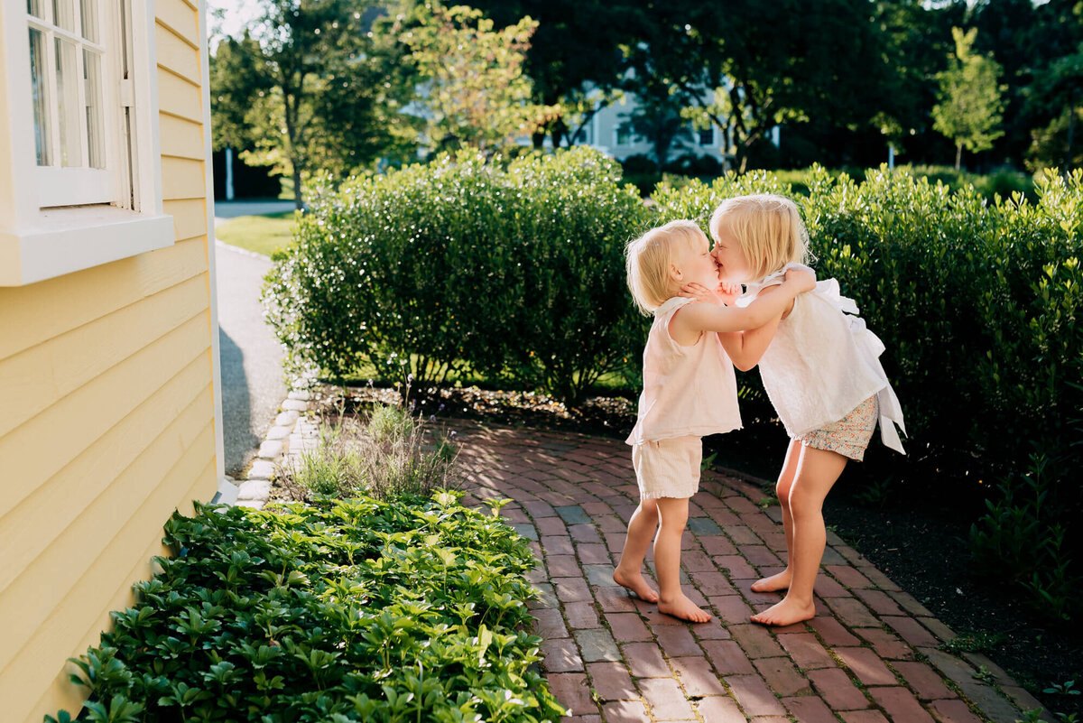 Two young sisters give each other a kiss on a brick path next to their house