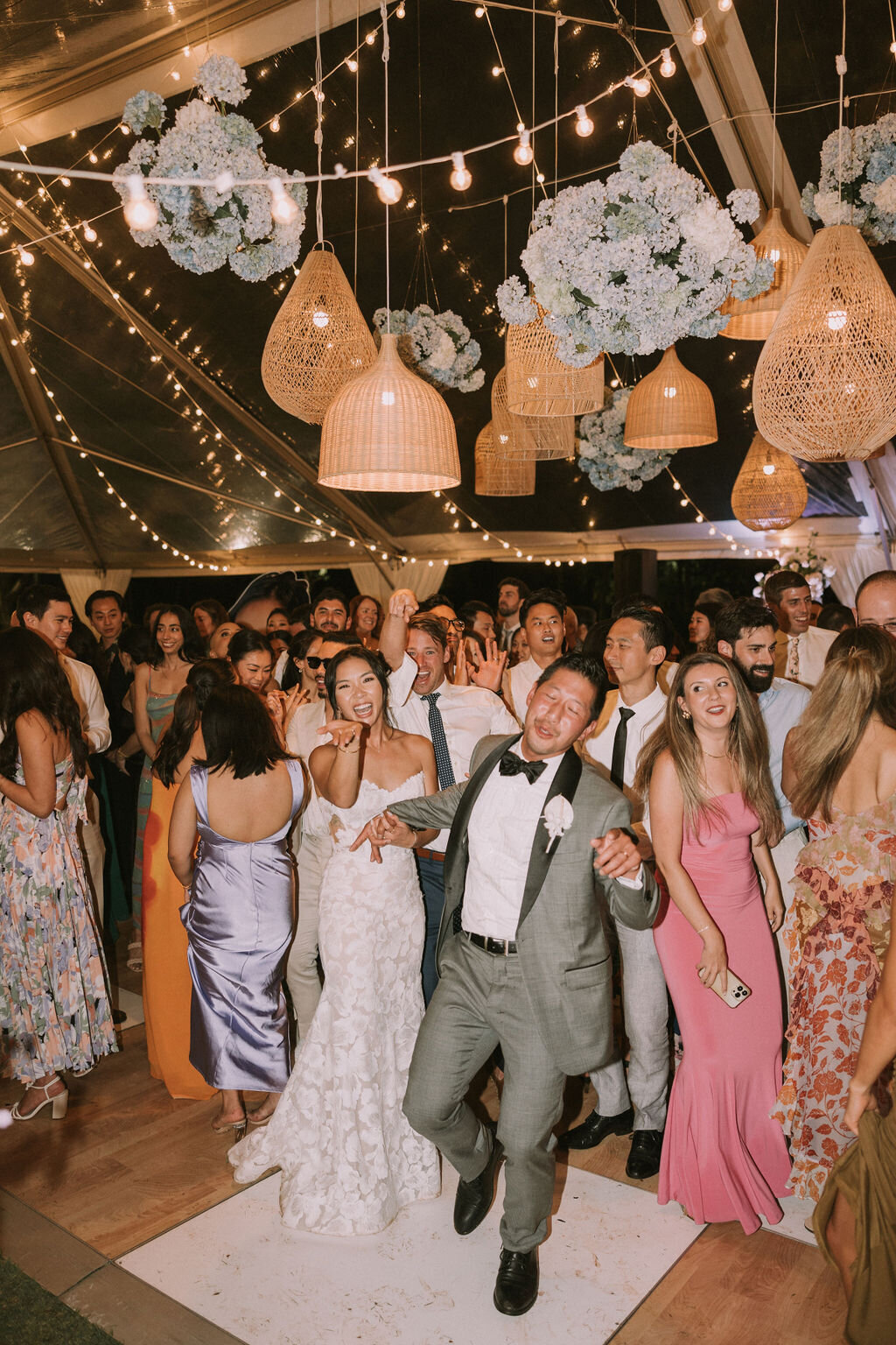 wedding couple posing silly towards camera while their guests begin dancing under clear tent and blue and white hydrangea and lantern installation
