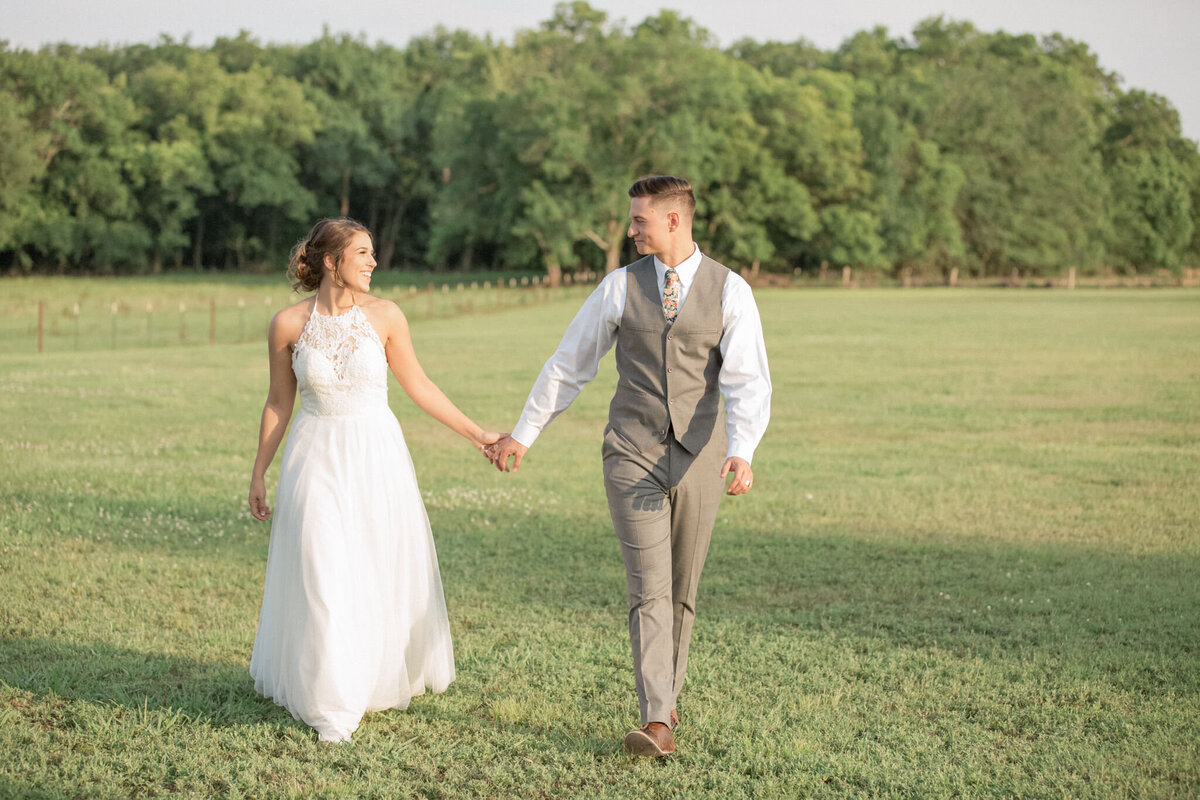 The new couple walk toward the camera in an open field, looking at each other and holding hands