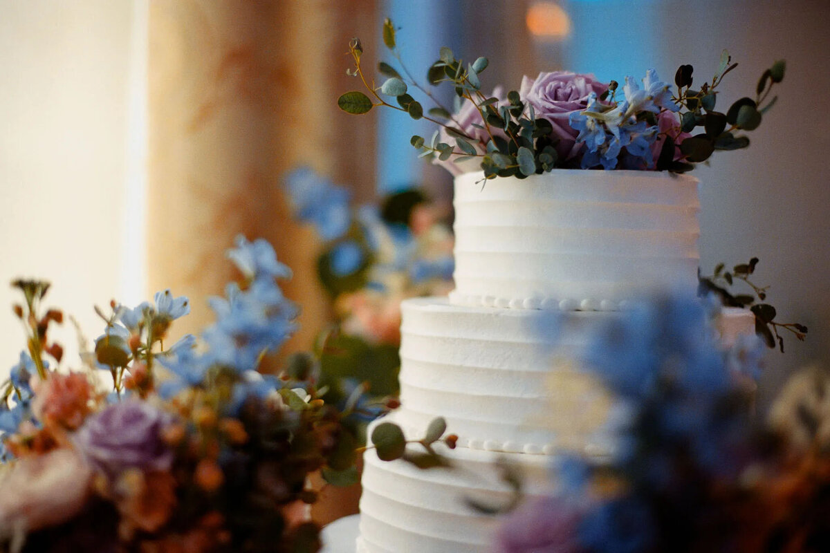 A three-tiered white wedding cake decorated with pink and purple flowers and greenery, surrounded by matching floral arrangements in soft focus—captured beautifully by an NJ wedding photographer.