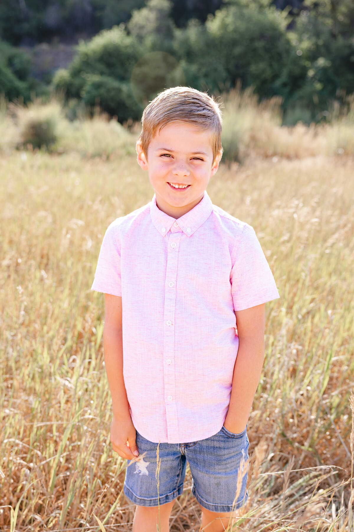 A young boy with short blonde hair stands confidently in a golden field, wearing a light pink button-up shirt and denim shorts, smiling under the warm Midway, Utah sun.