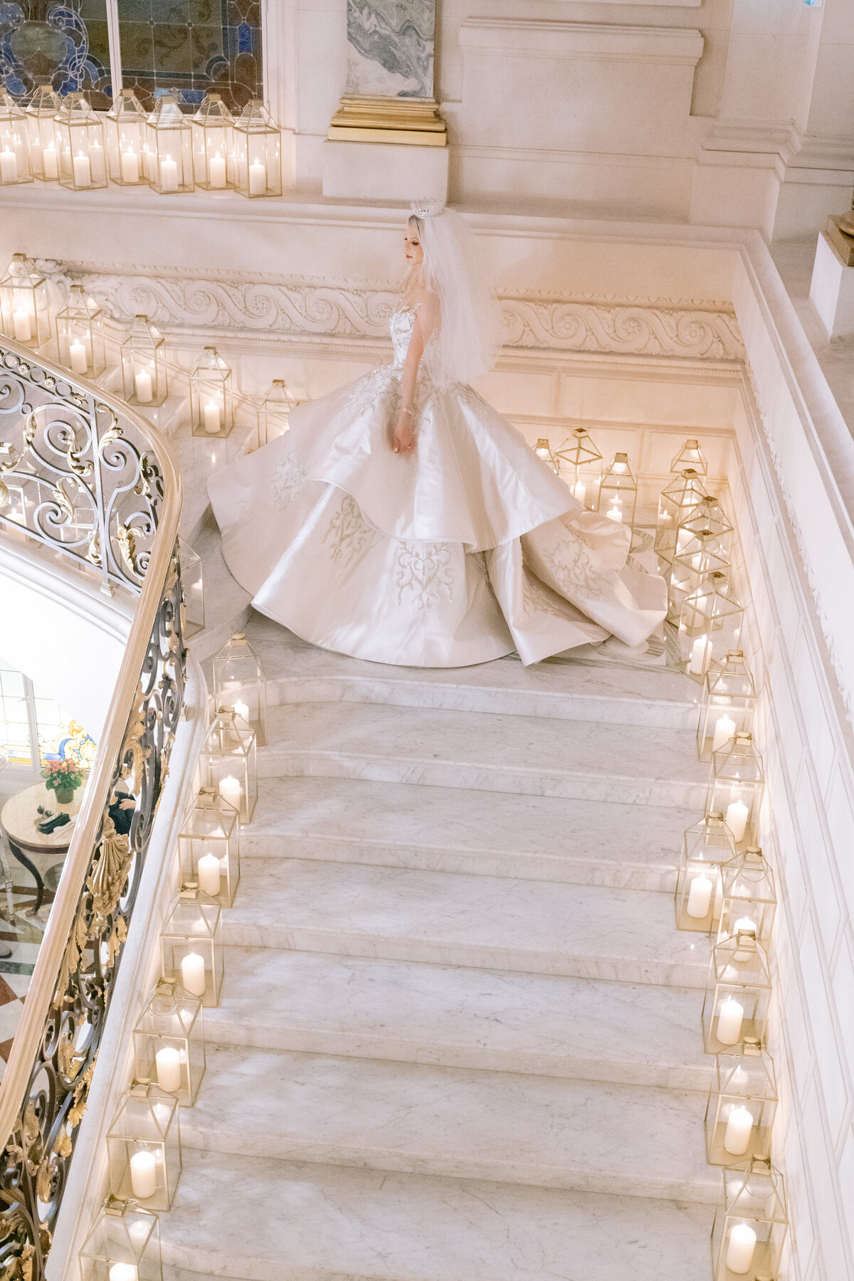 The beautiful photo was taken at two iconic locations, the Shangri La Hotel and the Ritz Paris Hotel in Paris, France, by the talented photographer Matteo Coltro. In the enchanting scene captured, a radiant bride stands elegantly on a grand marble staircase adorned with numerous candles in glass lanterns. She exudes grace in a voluminous white wedding dress with intricate embroidery and a veil, her gaze turned away from the camera. The staircase is further enhanced by ornate railings, adding to the grandeur of the setting. Additionally, Zen Film Works provided videography services to capture this magical moment on film.