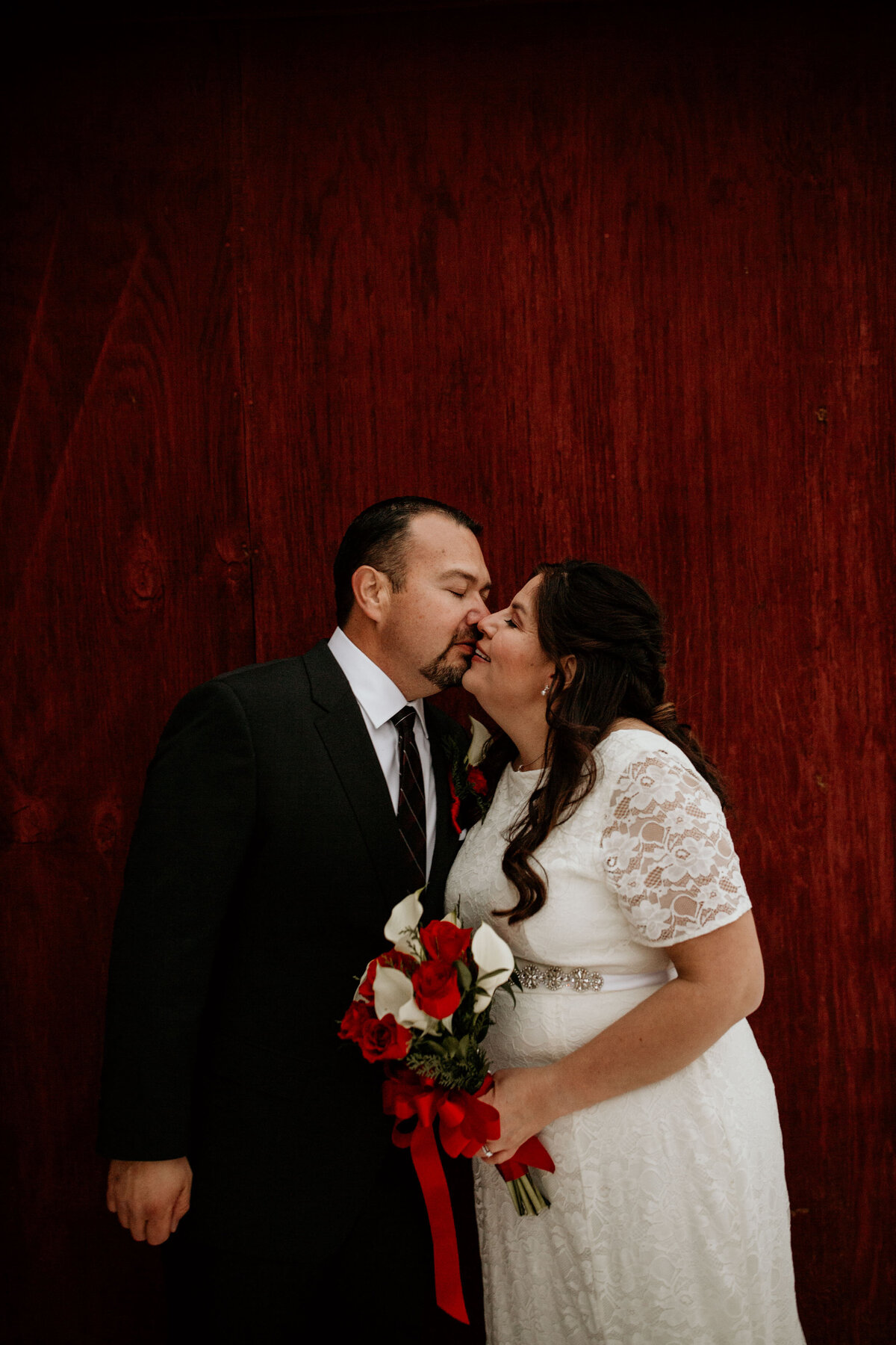 groom kissing bride on cheek in front of a red barn wall
