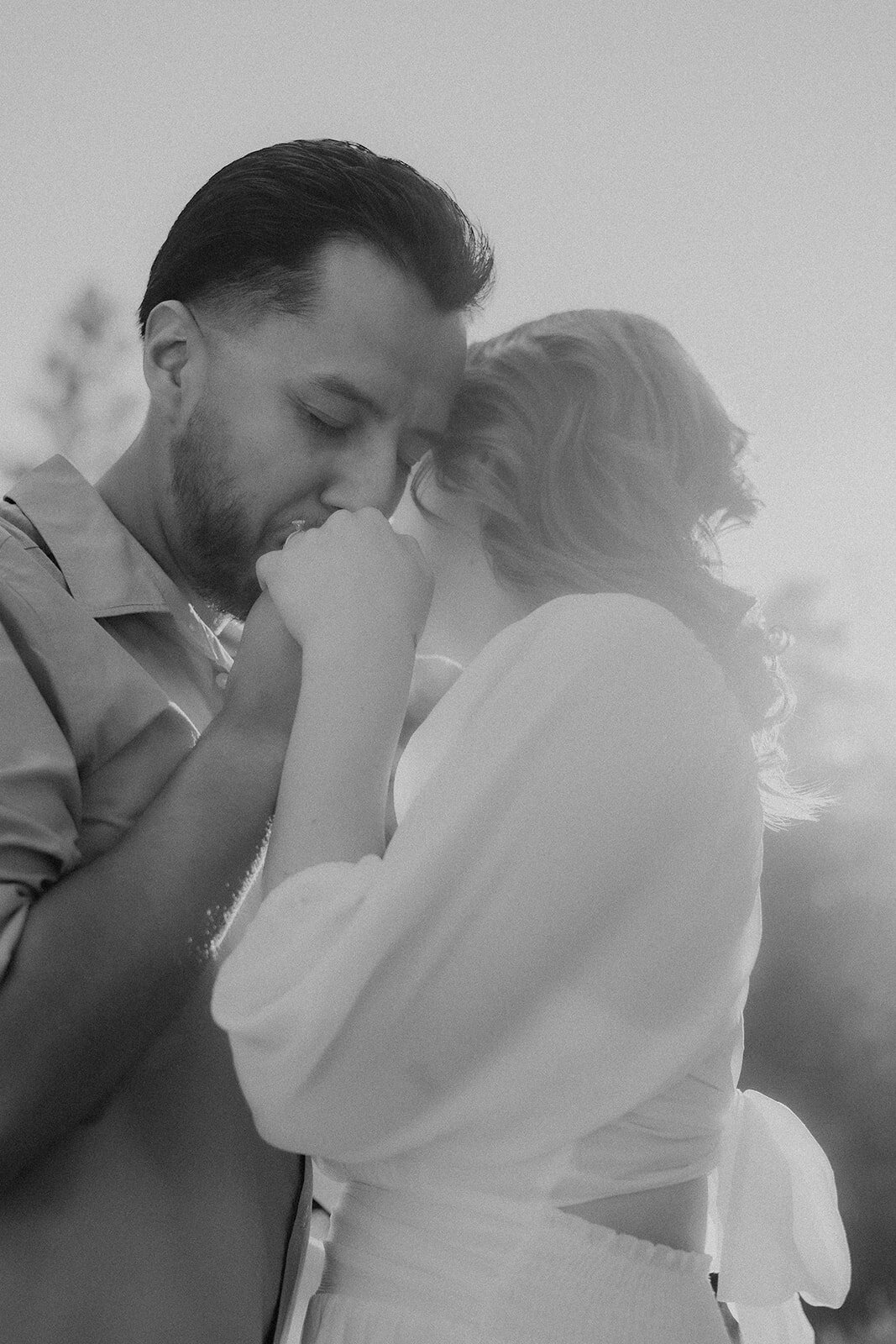 A tender black-and-white moment of the couple, with the man gently kissing the woman’s hand while they embrace.
