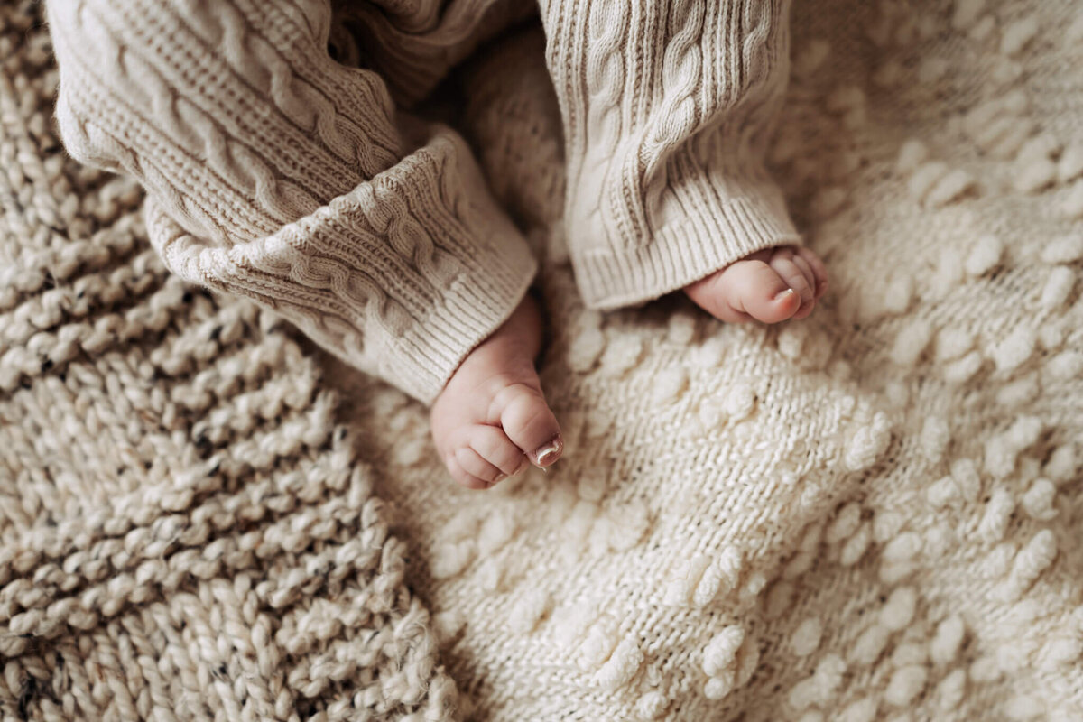 A close-up of a baby's feet resting on a textured cream-colored knit blanket. Perfect for portraits or a milestone celebration, the baby wears cozy cable-knit beige pants, and the soft patterns of the blanket add warmth to the scene.