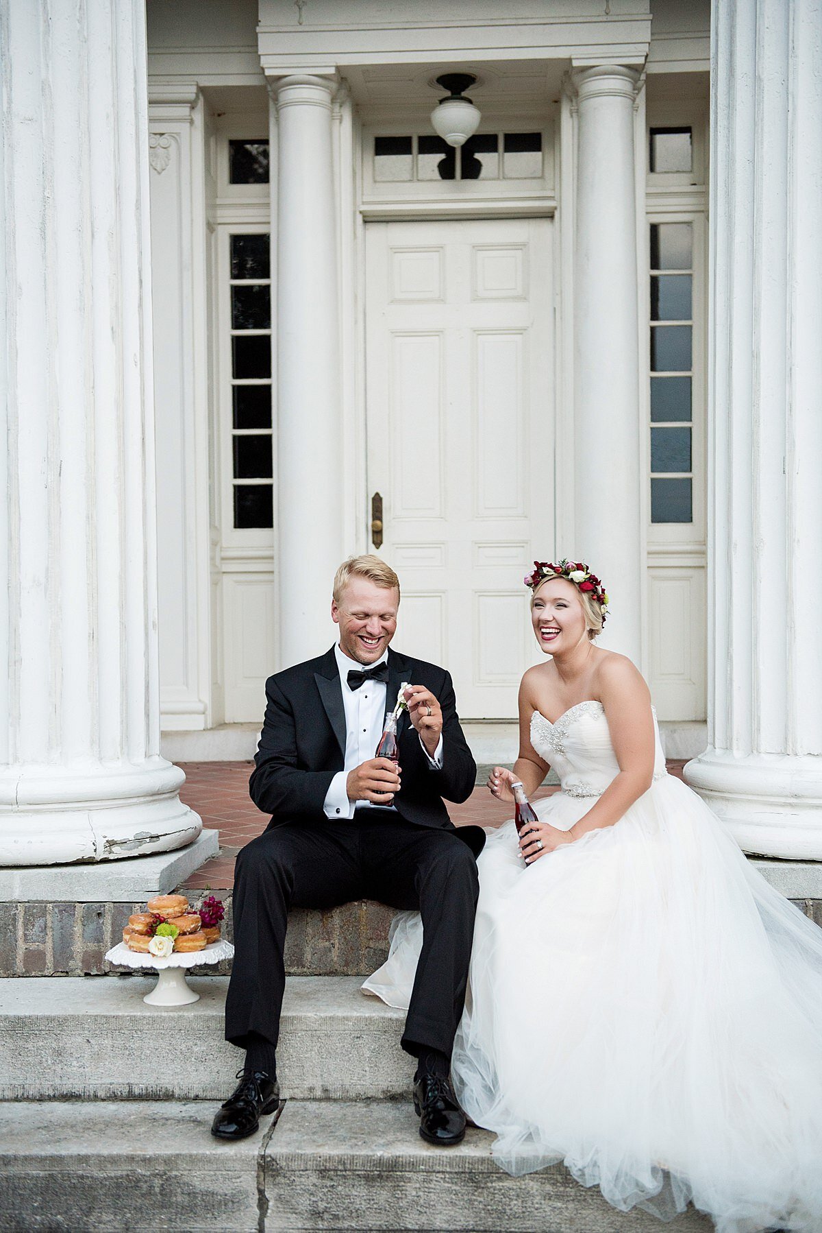 The bride and groom sit on the front steps of Rippavilla Mansion amongst the tall white columns. They are drinking Cheerwine from glass bottles next to a small two tier peach cake on a white pedestal cake stand. The groom is wearing a black tuxedo with a white shirt and black bow tie. The bride is wearing a flower crown with white and red flowers and a strapless dress with a fitted bodice and sweetheart neckline. The tulle skirt of her dress is spread out over the stone steps.