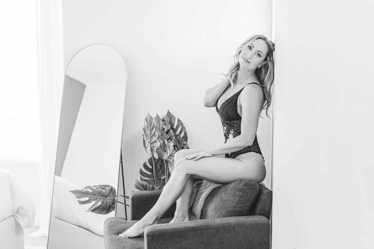 A woman sitting on a chair in black lingerie, posing confidently near a mirror and surrounded by greenery.