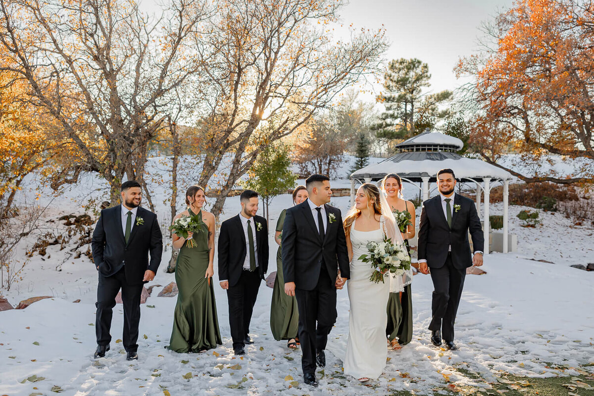Bride and groom and bridal party walking hand in hand on snow with breathtaking Colorado Springs fall leaves view, captured by Michelle Betz Photography, a Colorado Springs wedding photography expert.