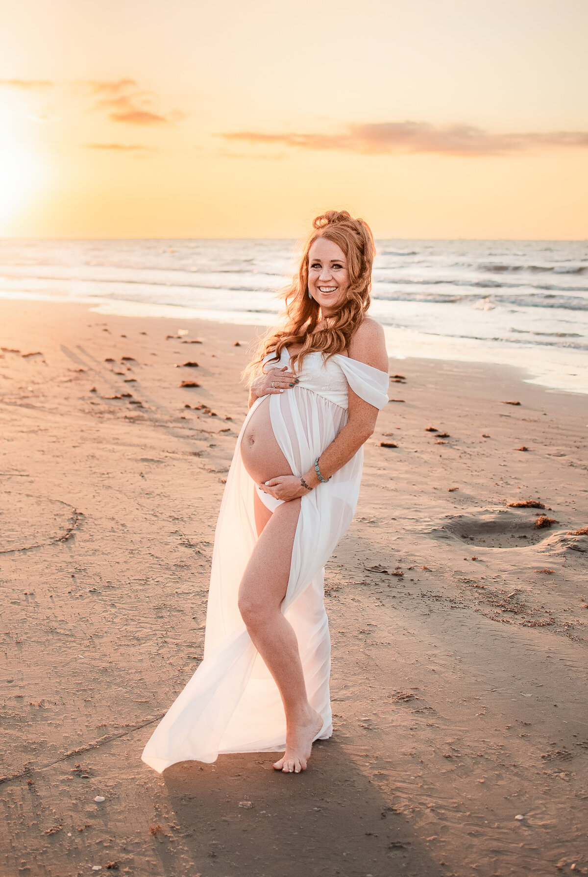A pregnant mom wearing a white dress stands on a Galveston beach at sunrise.
