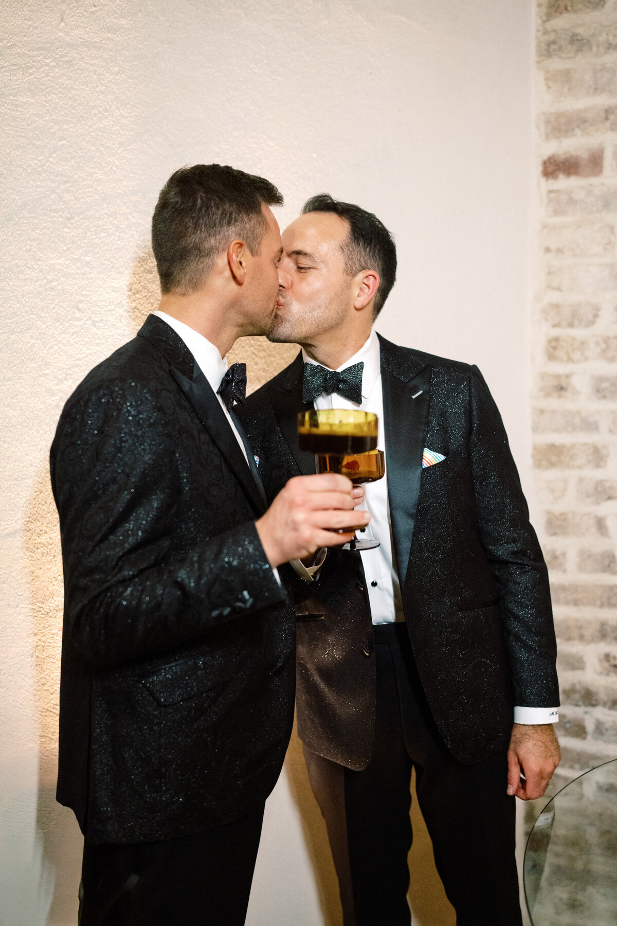 Two men elegantly dressed share a tender kiss, holding drinks, capturing a moment of love and connection amid a luxurious setting, showcasing the beauty of affection at a celebration.