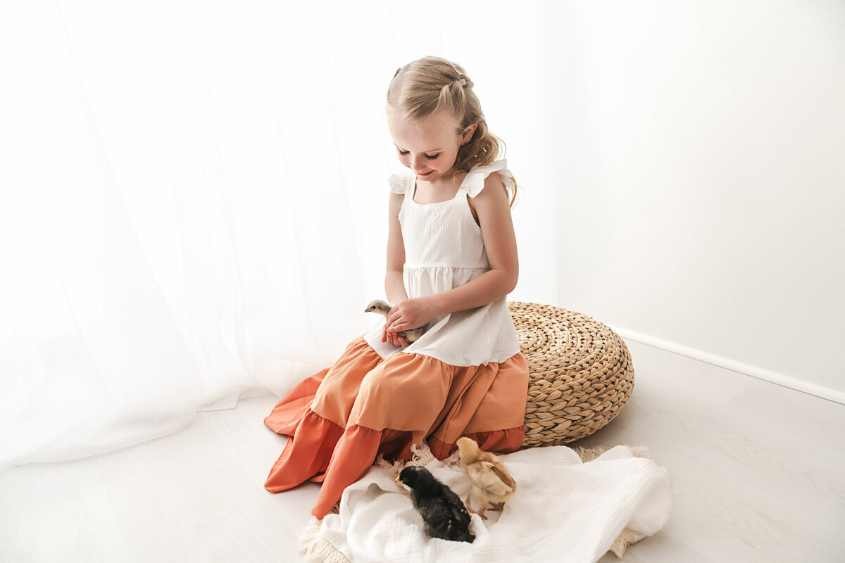 A young girl in a white dress sits on a pouf, engrossed in play with her baby chicks laid out on a soft white blanket, in a bright, airy room.