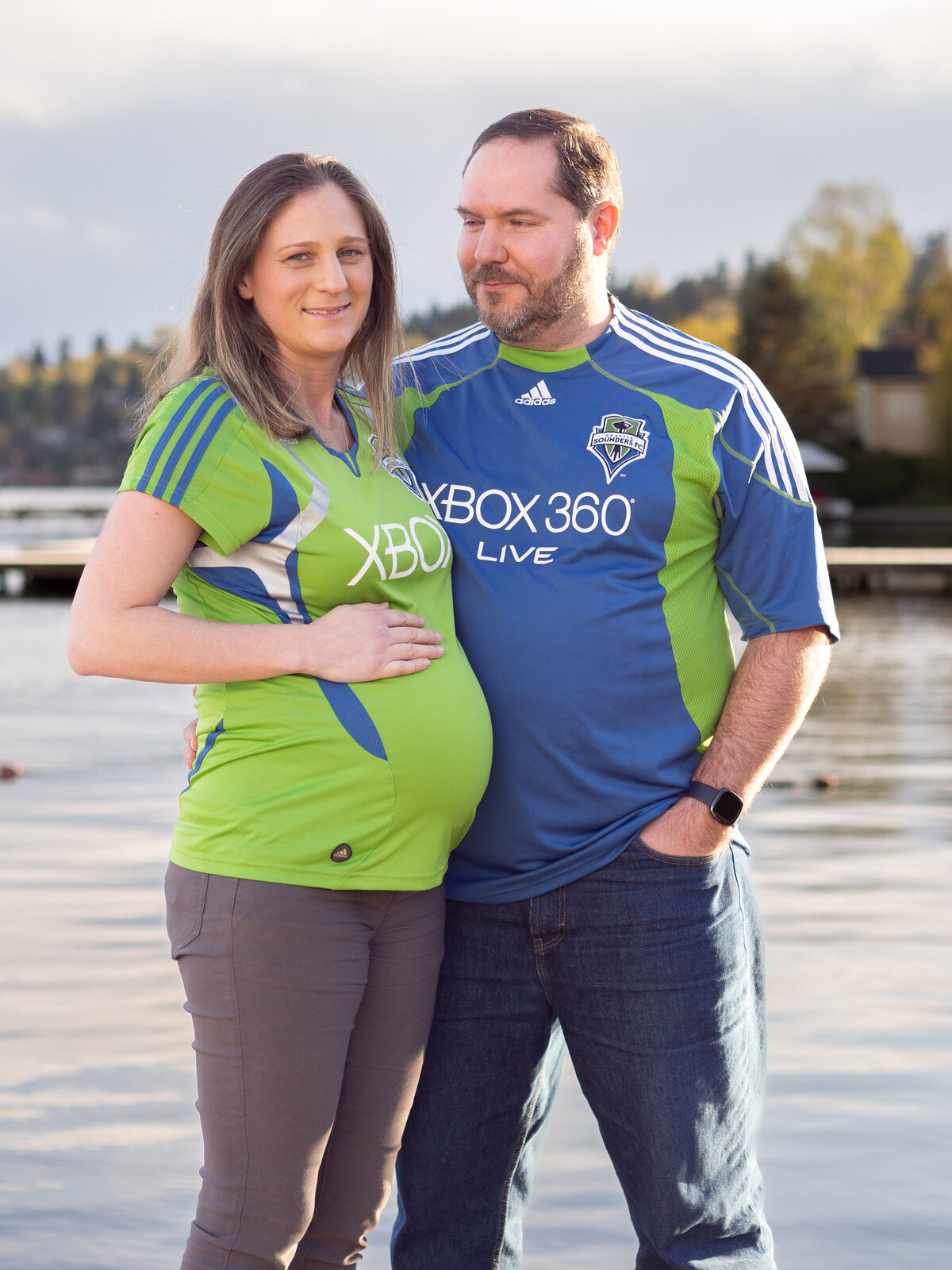 a couple standing by Lake Washington in Lake Forest Park smile excitedly as they anticipate the birth of their new baby.