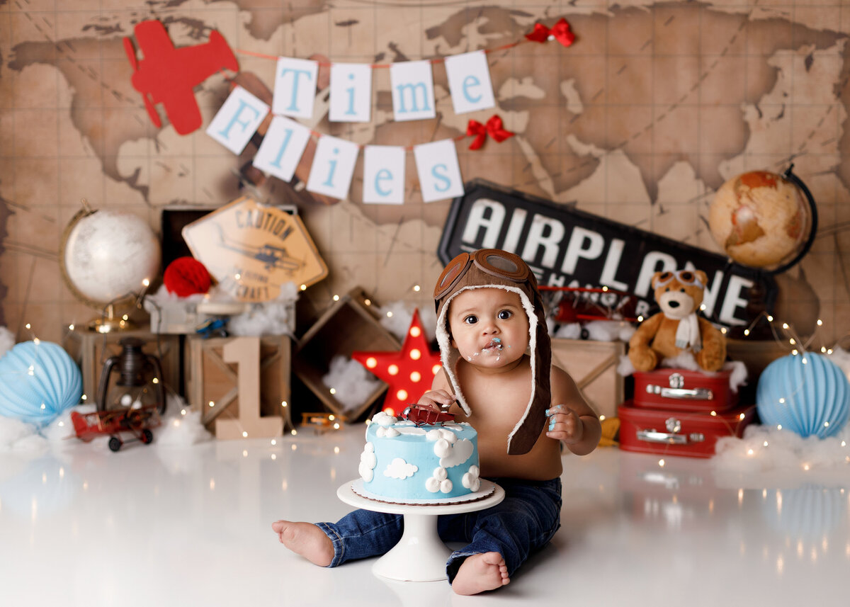 Airplane pilot themed cake smash. Baby boy wearing vintage pilot cap and jeans, sitting in front of a blue cake with white clouds and a vintage red airplane on the top. Baby has as bit of blue icing on his face. In the background, there is a vintage world map, vintage suitcases, globes, and airplanes.