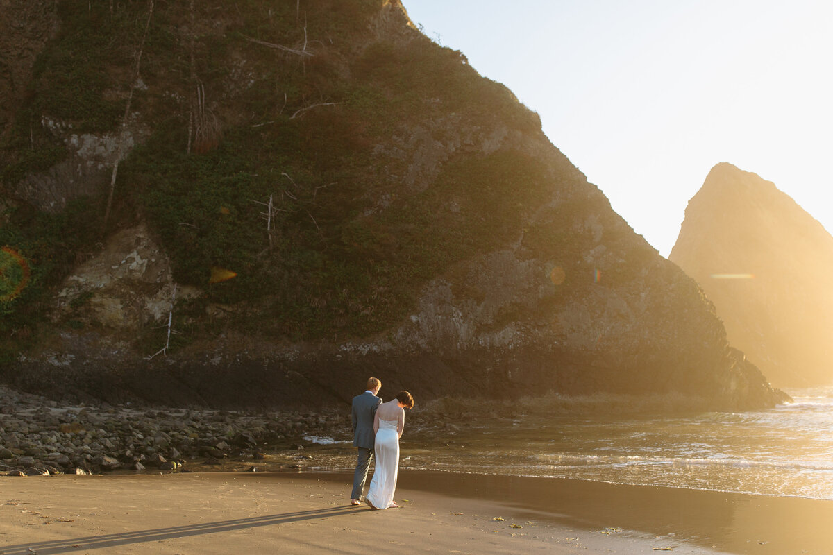Wide shot of bride and groom at Arch Cape walking into the water.