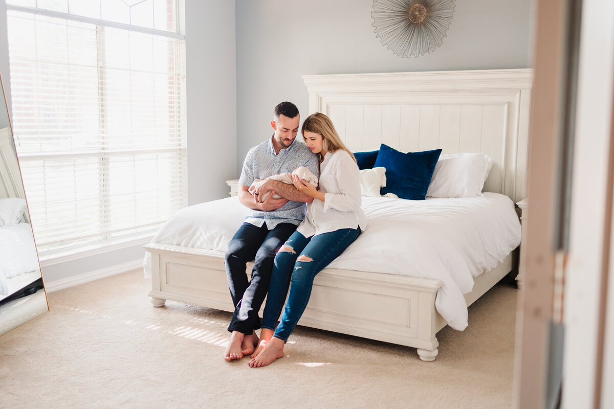 Parents hold a newborn baby girl, they are sitting on the bed with white bedspread and dark blue cushions. The woman with blonde hair is wearing a white jean blouse and the man is wearing a gray t-shirt