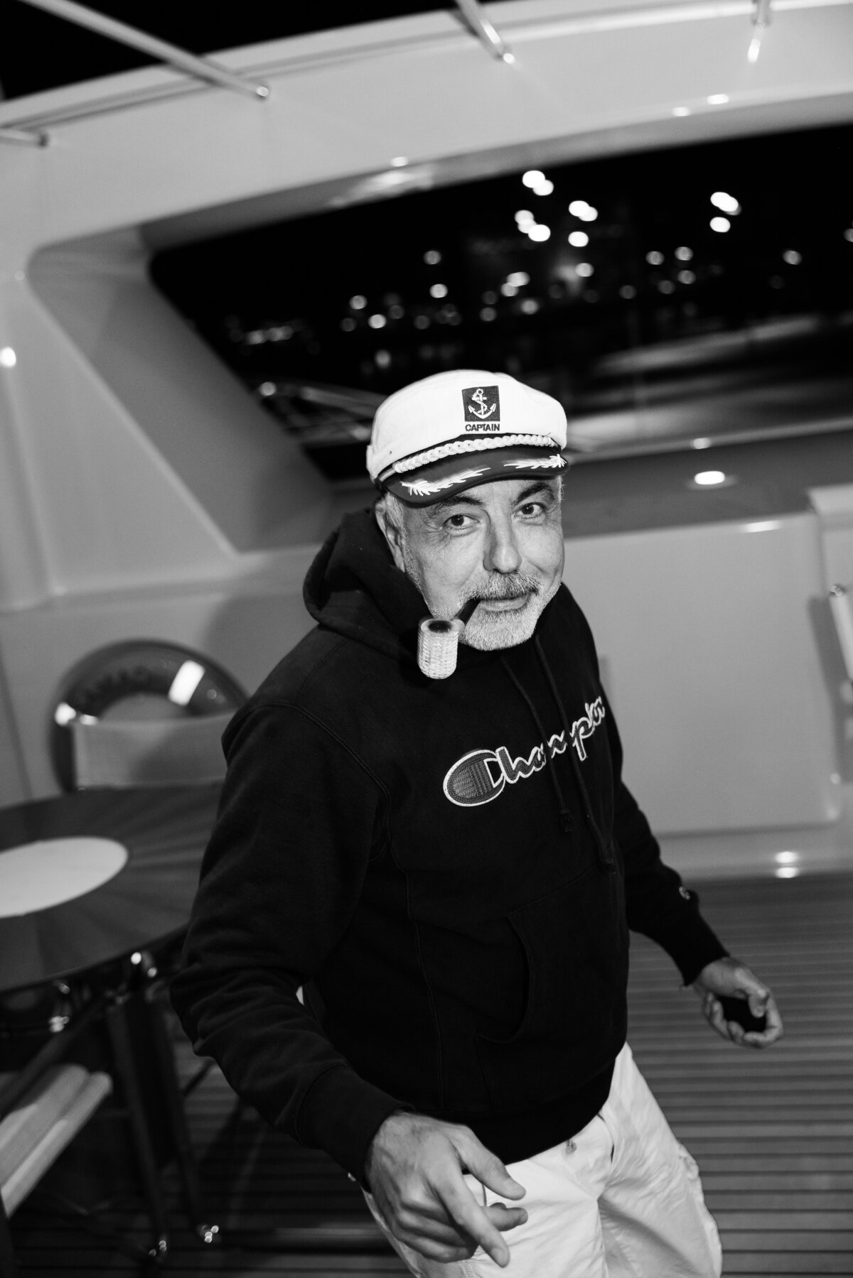 A black-and-white portrait of a man wearing a captain's hat and smoking a pipe on a luxury yacht in Amsterdam. This image captures the relaxed, high-end lifestyle atmosphere of the event.