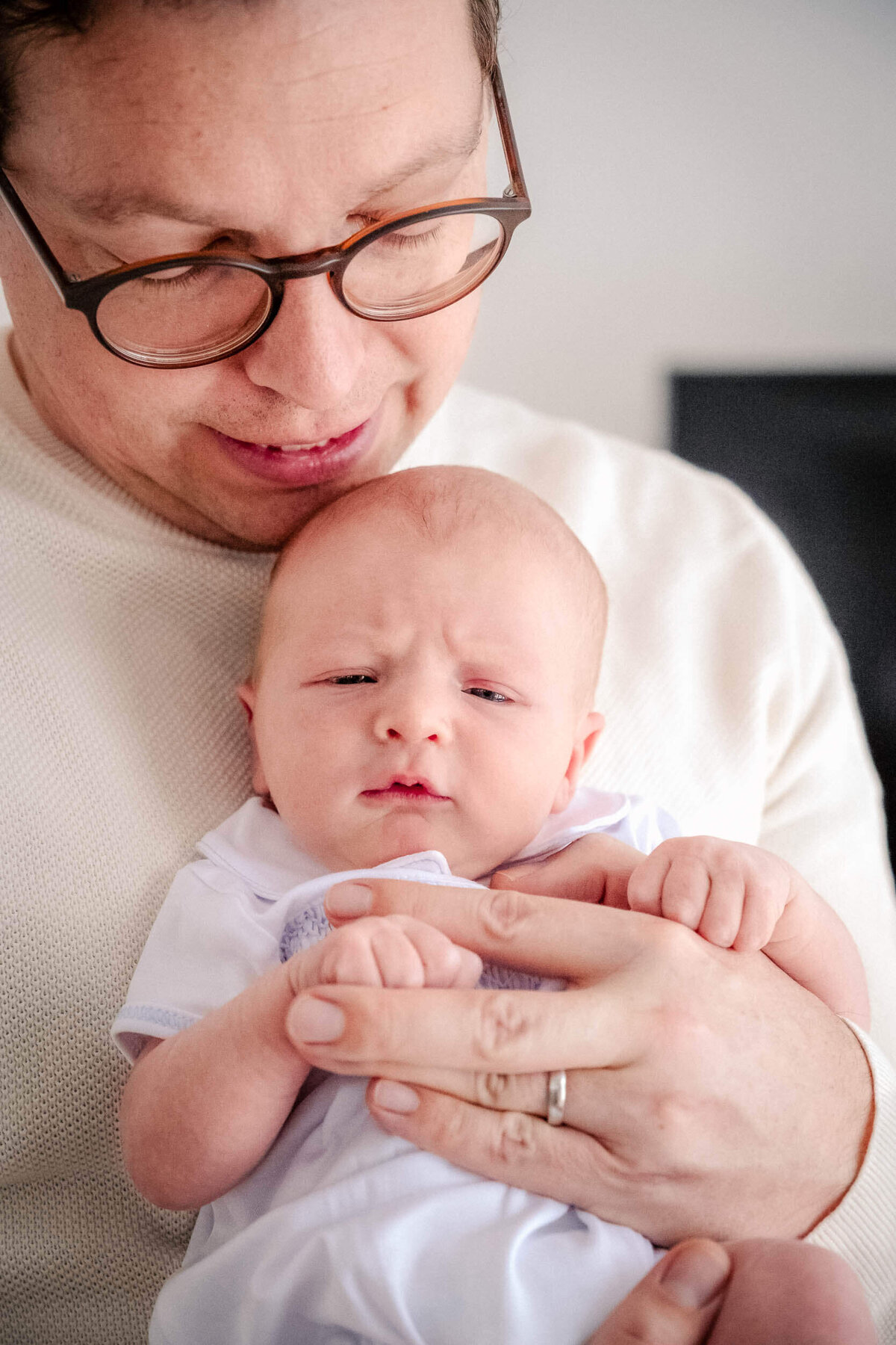 Father in glasses holding a newborn baby close to his chest, both looking softly toward the camera.