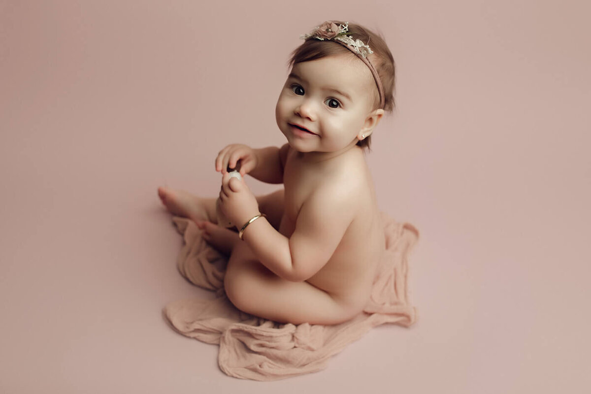 a nude one year old girl sitting on a light pink backdrop wearing a bracelet