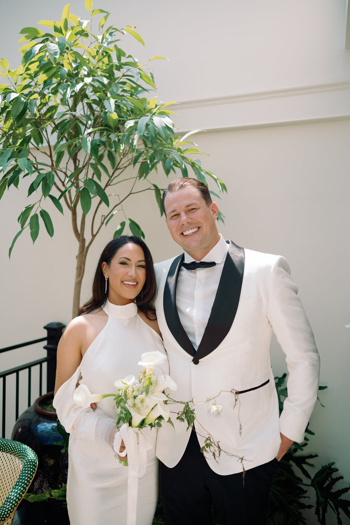 Bride and groom smile warmly while holding a bouquet, standing together in their chic wedding attire.