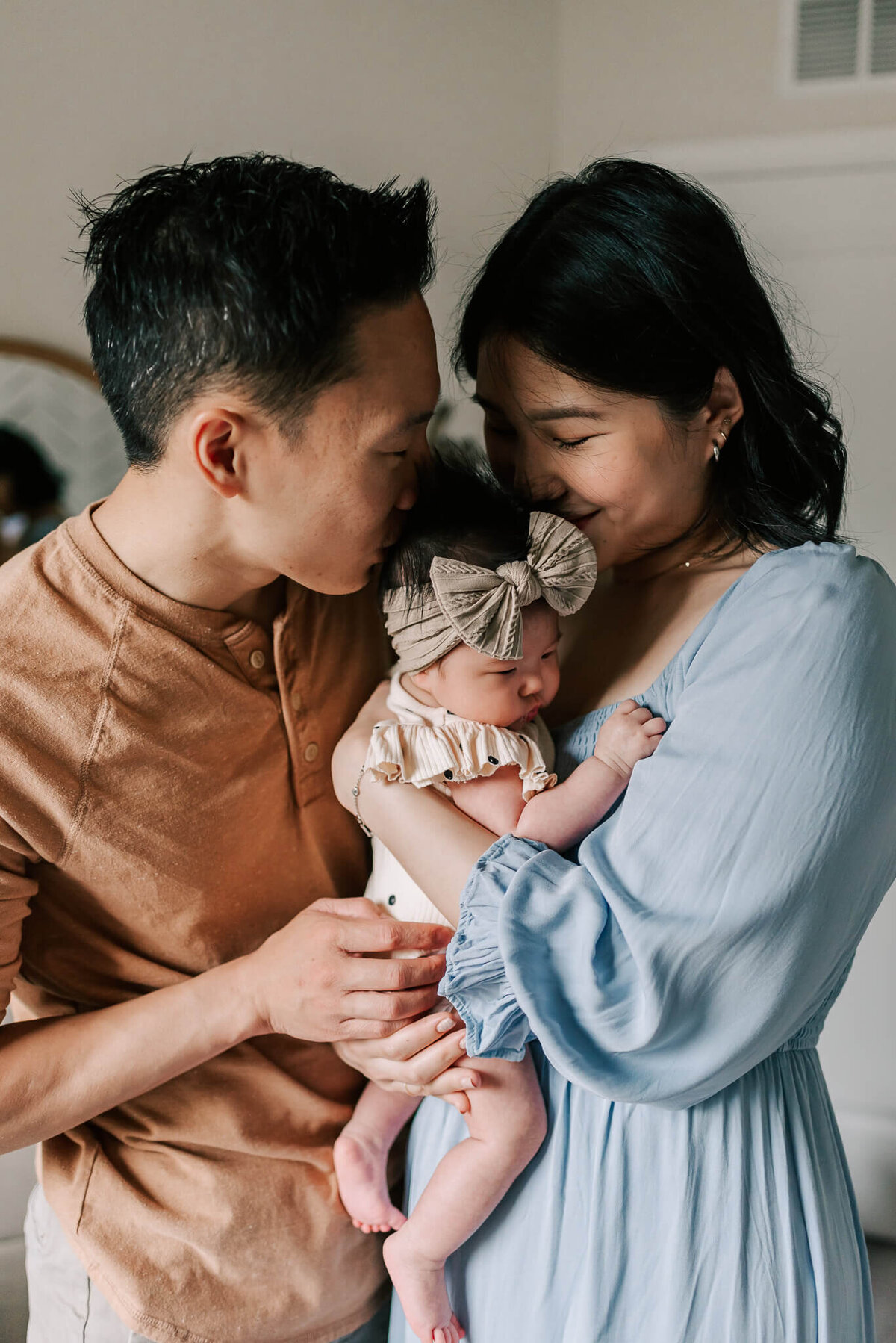 a newborn being snuggled by her parents, the mom in dusty blue and father in muted brown