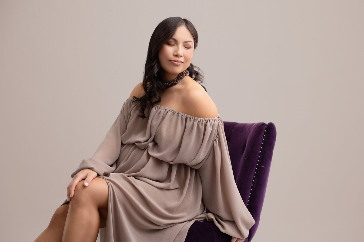 An elegant image of a pregnant mother sitting on a deep purple chair and wearing a light brown off the shoulder gown.
