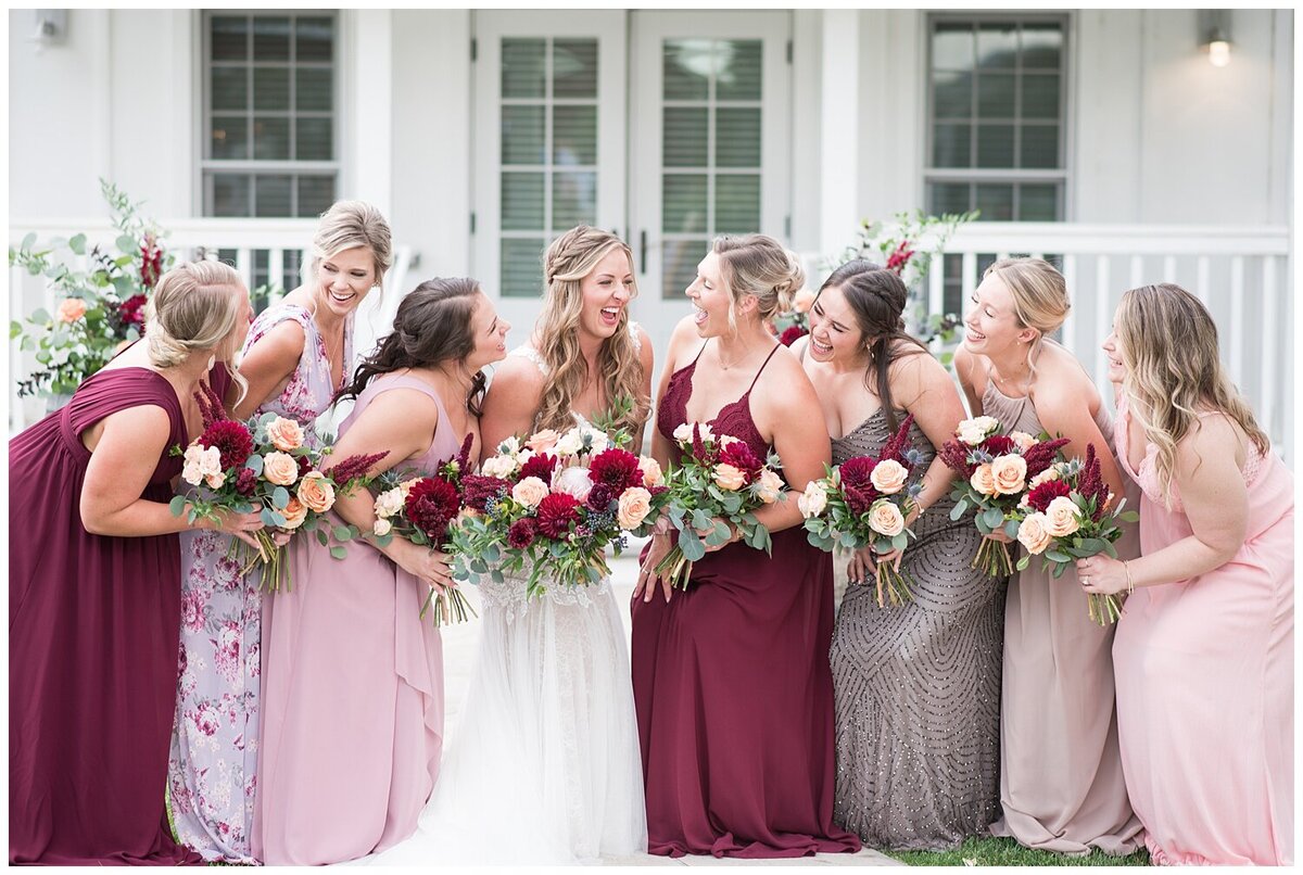 At Barn at Raccoon creek, a Denver wedding photographer captures a bride in her wedding dress standing with her bridesmaids in all different shades of red and pink dresses with their flowers held up at their sides and smile at each other