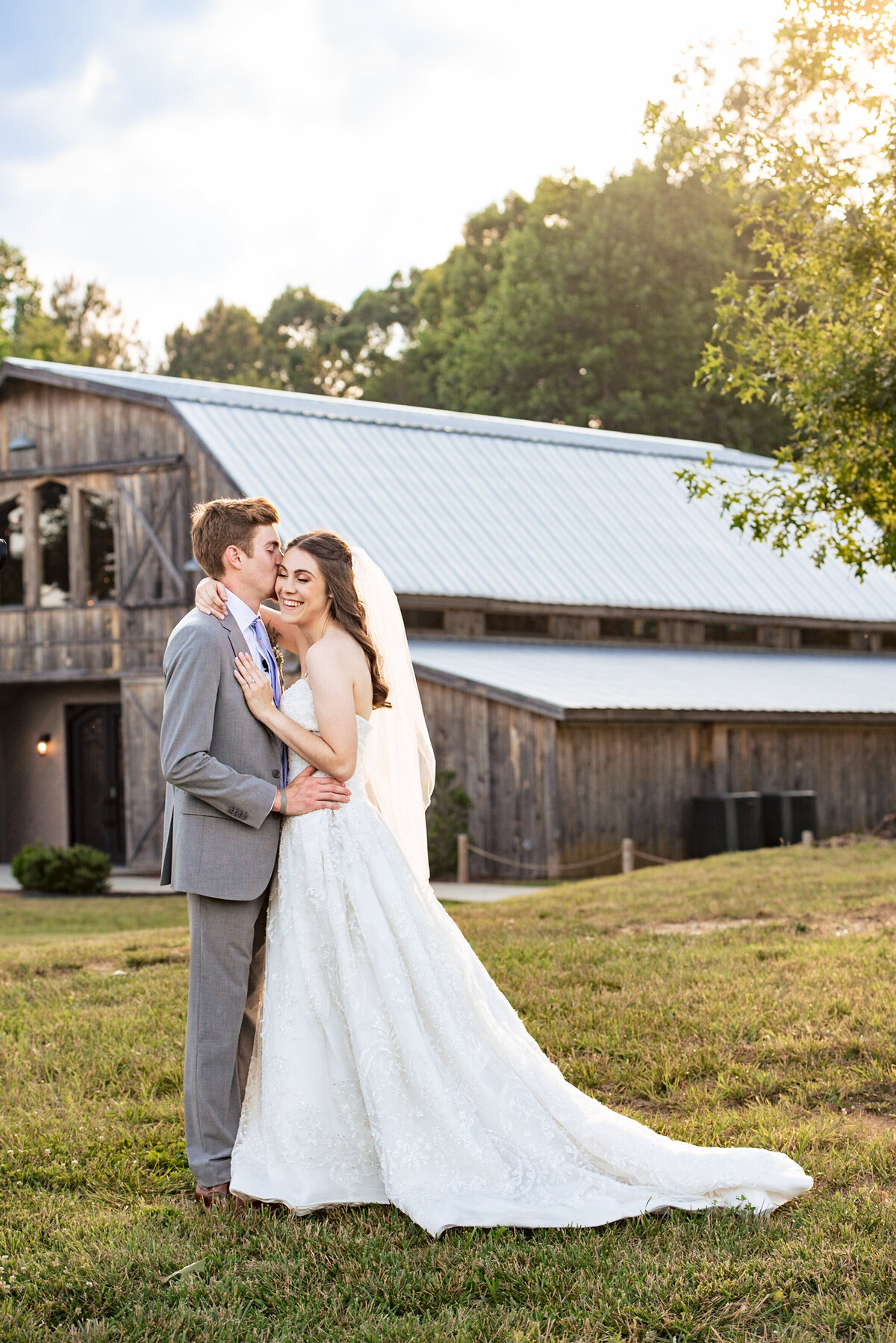 Outdoor portrait of bride giggling as groom kisses her temple during sunset showing the barn in the background at Lady Bird Farms by Charlotte Wedding Photographers DeLong Photography