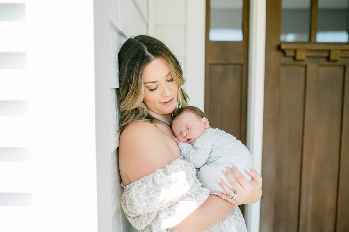 A mom leans against the side of her house while holding her newborn baby boy on her chest