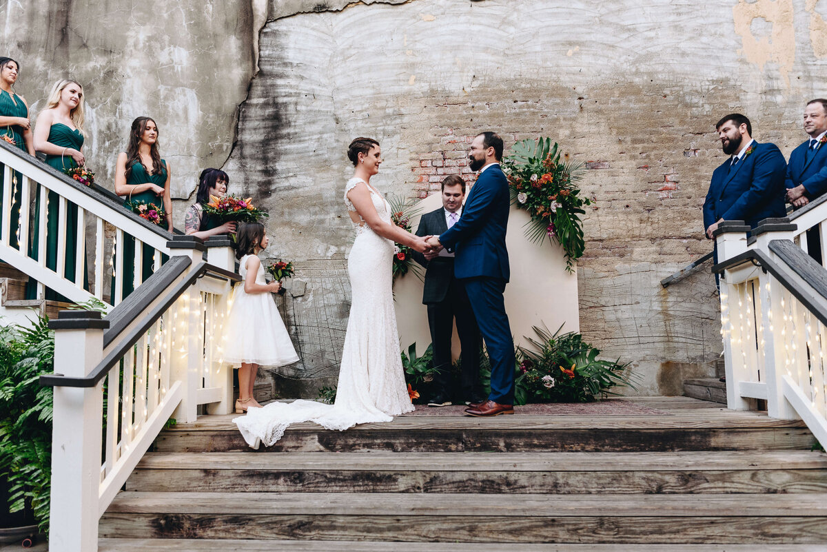A bride and groom stand hand-in-hand at the center of an outdoor wedding ceremony, flanked by bridesmaids in green dresses and groomsmen in blue suits on the stairs, with an officiant and flower girl nearby.