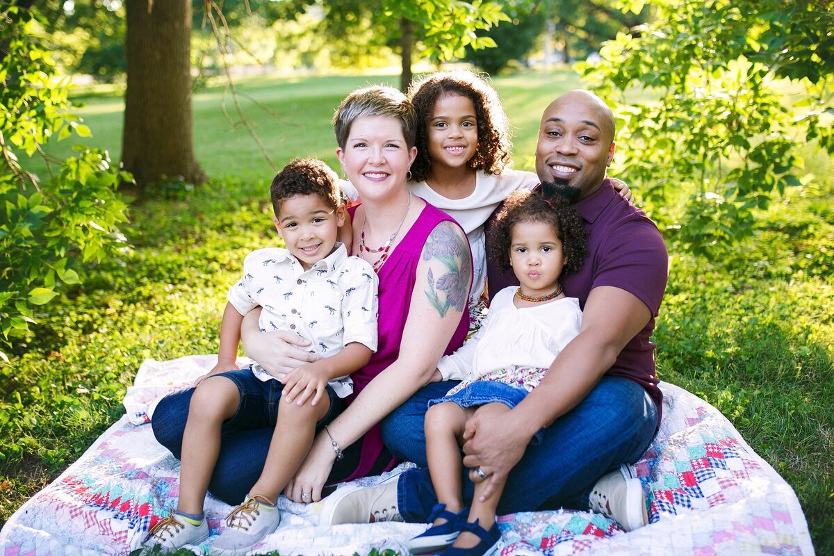 A-smiling-family-sitting-on-a-quilt-in-a-scenic-green-space-in-the-Meridian-Kessler-neighborhood-on-the-north-side-of-Indianapolis-during-a-sunny-family-photo-session-surrounded-by-lush-trees-and-nature