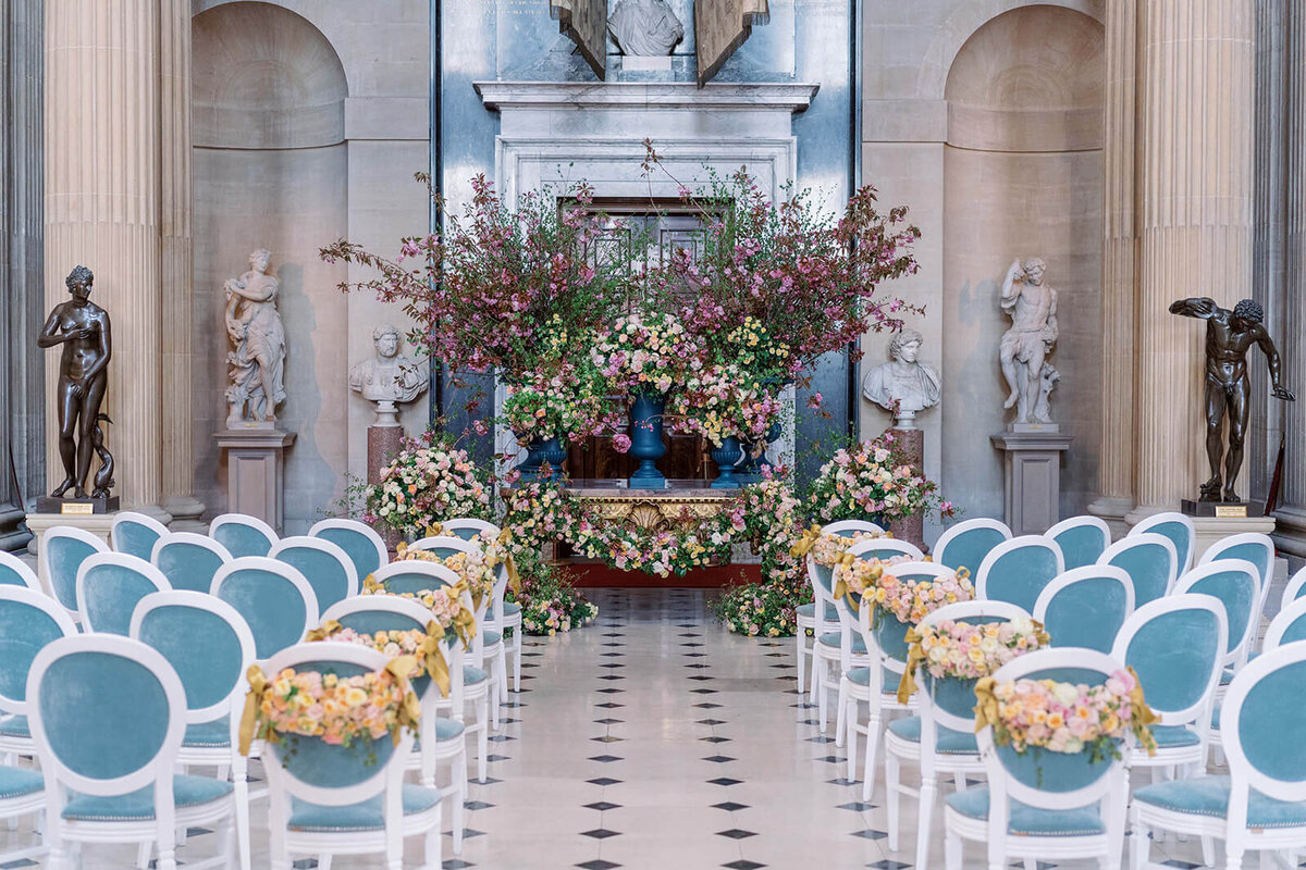 view looking down a wedding aisle at blenheim palace with spring blossom trees at the end of the aisle and blue chairs lining the aisle with floral swags